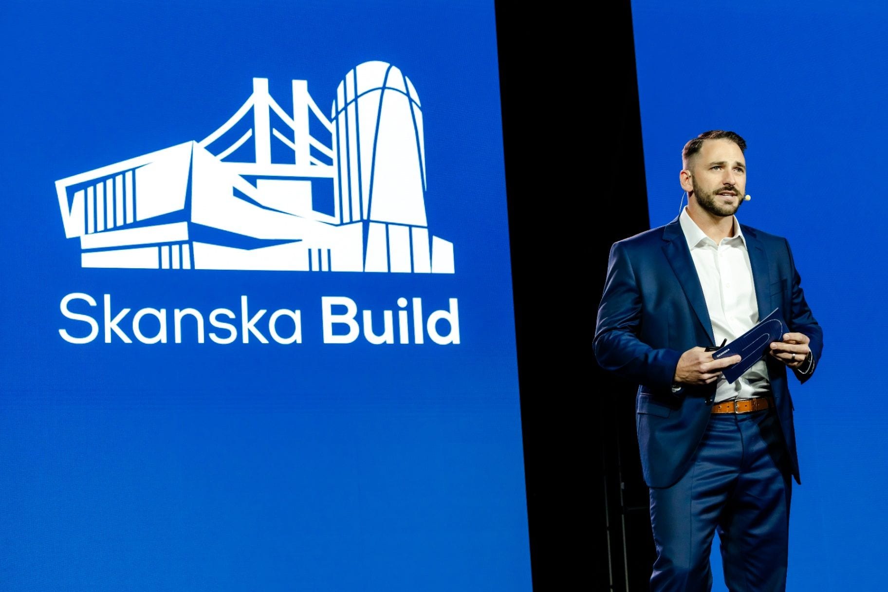 A corporate speaker presenting on stage beside a Skanska Build presentation screen